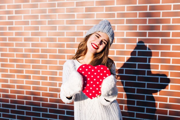 Portrait jolie fille aux cheveux longs en bonnet tricoté, pull chaud sur le mur extérieur. Elle tient un grand coeur rouge dans des gants, souriant.