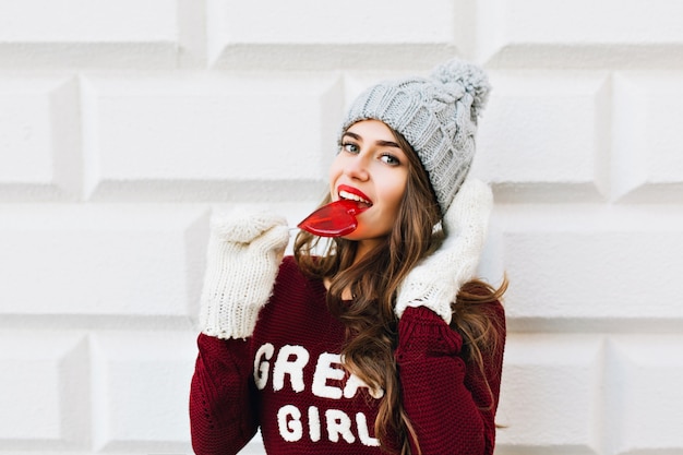 Portrait jolie fille aux cheveux longs en pull marsala léchant sucette coeur rouge sur mur gris. Elle porte des gants blancs, souriant.
