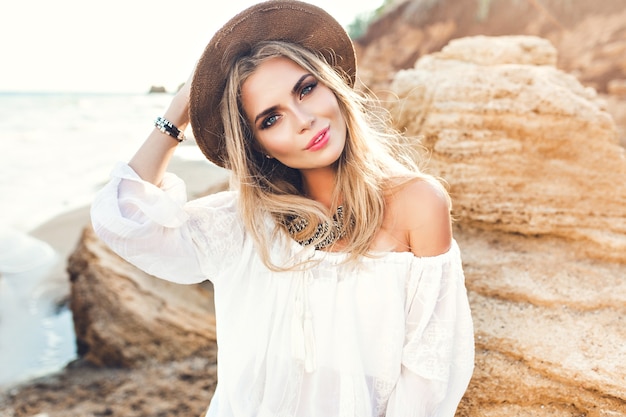 Portrait de jolie fille blonde aux cheveux longs posant sur une plage déserte. Elle sourit à la caméra.