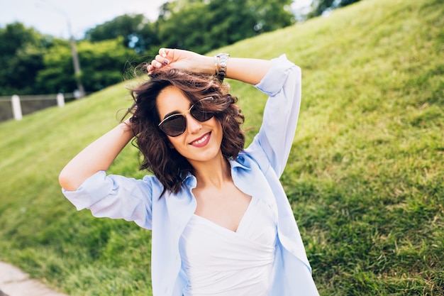 Portrait de jolie fille brune aux cheveux courts à lunettes de soleil souriant à la caméra dans le parc sur fond de pré. Elle porte un T-shirt blanc, une chemise bleue.