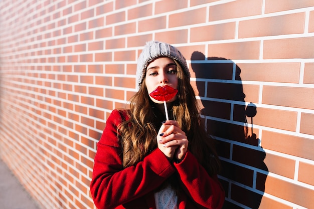 Portrait jolie fille brune avec des lèvres de sucette sur le mur extérieur. Elle porte un bonnet tricoté, un manteau rouge.