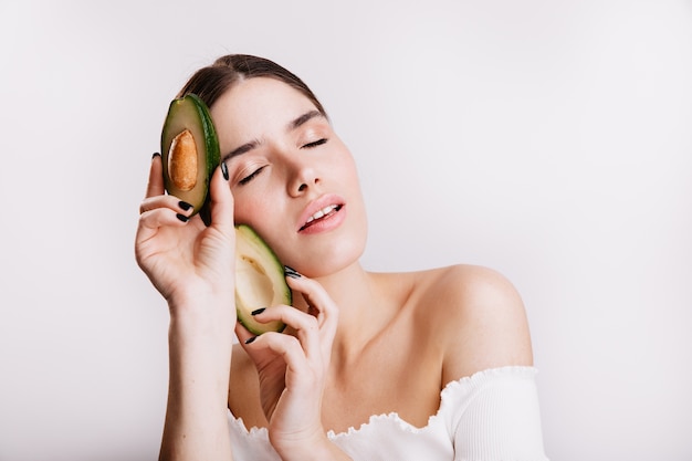 Portrait de jolie fille brune avec une peau claire et propre. Femme posant avec les yeux fermés tenant avocat sur un mur blanc.