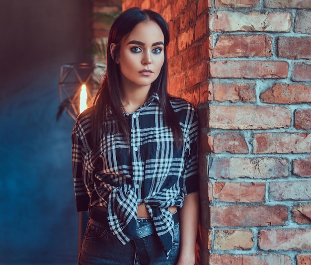 Portrait d'une jolie fille brune vêtue d'une chemise en flanelle et d'un jean s'appuyant sur le mur dans une pièce avec un intérieur loft.