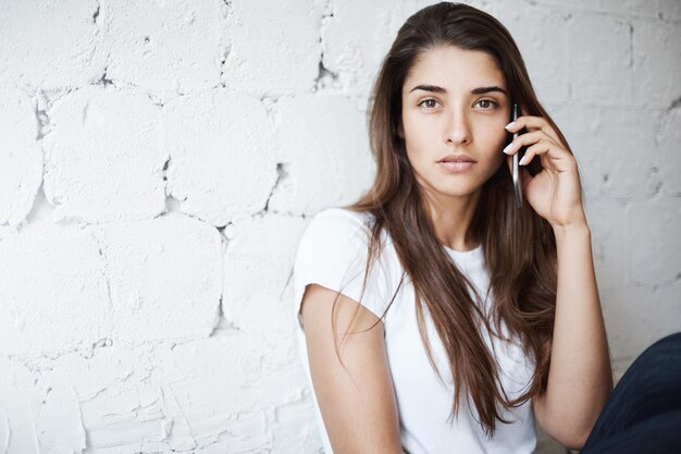 Portrait de jolie fille caucasienne, s'appuyant sur un mur de briques blanches tout en parlant via smartphone regardant rêveusement la caméra. La femme écoute son amie qui raconte ses derniers potins qui se répandent dans la ville.