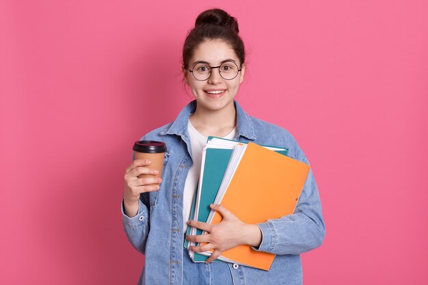 Portrait de jolie fille avec chignon en veste en jean et t-shirt blanc avec un doux sourire sur rose détient des dossiers sur rose