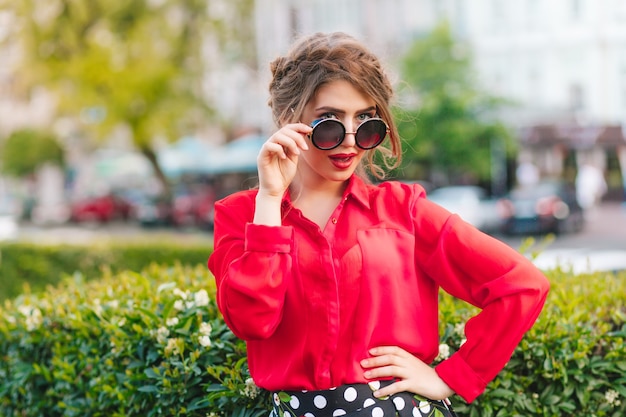 Portrait de jolie fille à lunettes de soleil posant à la caméra dans le parc. Elle a une coiffure, un chemisier rouge.