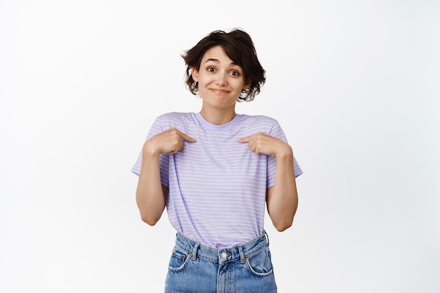 Portrait de jolie fille modeste pointant du doigt sur elle-même et souriant, c'est moi le geste debout sur blanc