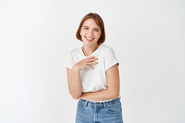 Portrait d'une jolie fille naturelle riant, touchant la poitrine et souriante heureuse, debout en t-shirt et jeans contre le mur blanc