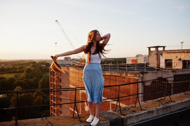 Portrait d'une jolie fille en vêtements décontractés posant sur le toit avec son chapeau à la main au coucher du soleil