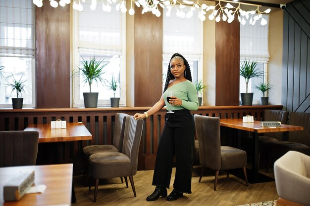 Portrait de jolie jeune femme afro-américaine portant un pull vert et un jean noir pose au restaurant avec un verre de vin rouge à portée de main