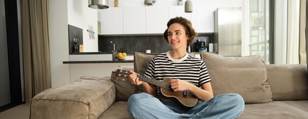 Photo gratuite portrait d'une jolie jeune femme assise sur un canapé qui apprend à jouer du ukulélé en tenant son instrument de musique