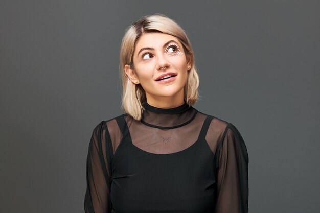 Portrait de jolie jeune femme blonde à la mode vêtue de vêtements élégants posant isolé en levant les yeux exprimant l'intérêt et la curiosité, ouvrant la bouche. Expressions faciales humaines