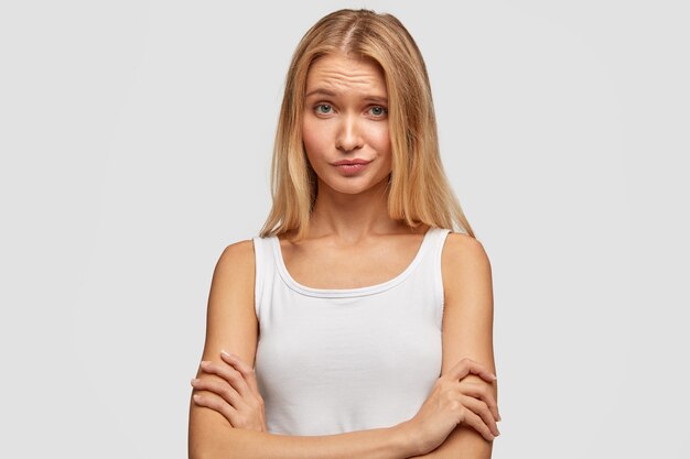 Portrait de jolie jeune femme avec une expression mécontente, garde les bras croisés, fronce les sourcils, se demande et entend quelque chose de désagréable, vêtu d'un gilet blanc, isolé sur le mur