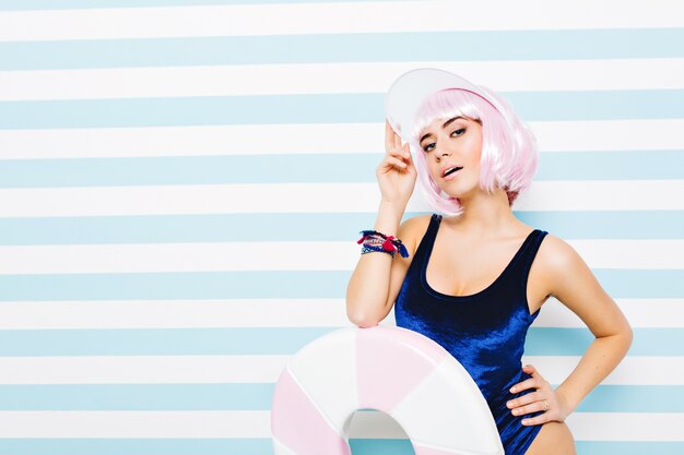 Portrait jolie jeune femme incroyable en body bleu reposant sur un mur rayé bleu-blanc. Porter une coiffure rose coupée, un bonnet de plage, une grosse sucette. Modèle sexy, humeur joyeuse.