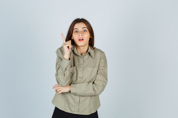 Portrait de jolie jeune femme pointant vers le haut, montrant le geste eurêka en chemise, jupe et regardant la vue de face intelligente