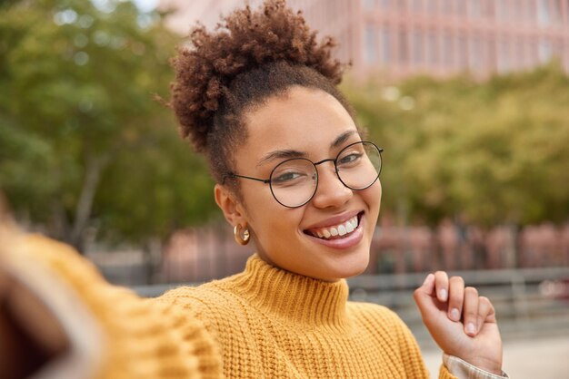 Portrait d'une jolie jeune femme souriante aux cheveux bouclés dans des lunettes rondes optiques pose pour des selfies à l'extérieur des promenades en milieu urbain pendant les loisirs photographie elle-même a une journée ou un week-end libre