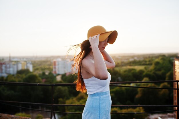 Portrait d'une jolie jeune femme en tshirt blanc et jupe bleue posant sur le toit avec son chapeau orange au coucher du soleil