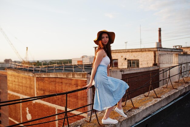 Portrait d'une jolie jeune femme en tshirt blanc et jupe bleue posant sur le toit avec son chapeau orange au coucher du soleil