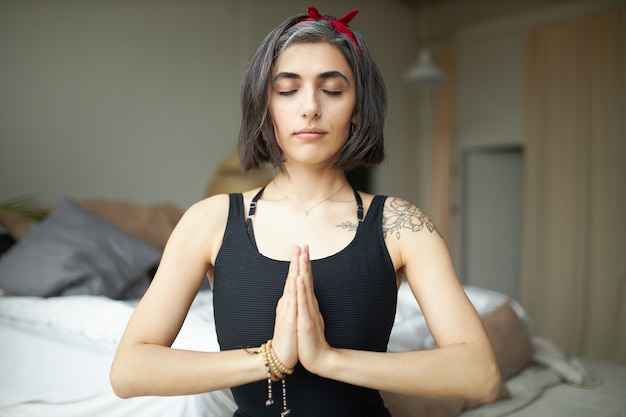 Photo gratuite portrait de jolie jeune femme yogi aux cheveux gris et tatouage assis en posture de lotus en gardant les mains pressées ensemble sur sa poitrine