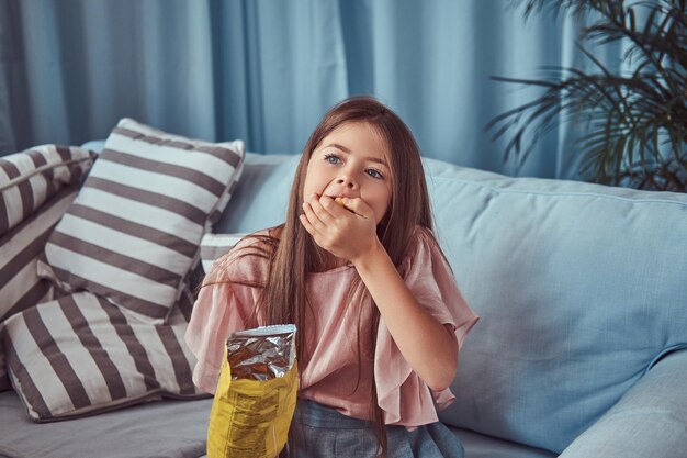 Portrait d'une jolie petite fille aux longs cheveux bruns, assise sur un canapé, mange des frites.