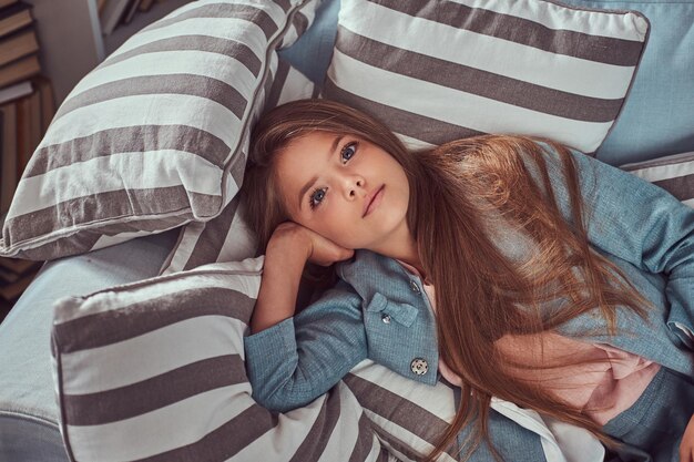 Portrait d'une jolie petite fille aux longs cheveux bruns et au regard perçant, regardant une caméra, allongée seule sur un canapé à la maison.