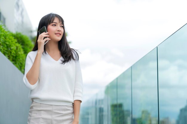 Portrait d'une jolie robe décontractée aux longs cheveux noirs asiatiques souriant à la main utilisant un smartphone avec un arrière-plan de paysage urbain et de ciel de lumière blanche liberté de fond et joyeux émotionnel