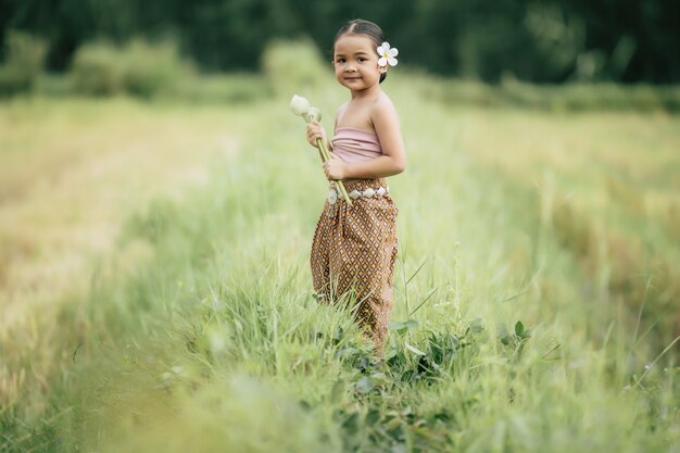 Portrait de jolies filles en costume traditionnel thaïlandais et mettant une fleur blanche sur son oreille, debout et tenant deux lotus à la main sur une rizière, elle sourit de bonheur, espace pour copie