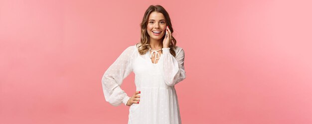 Portrait de joyeuse belle femme blonde en robe blanche parlant au téléphone tenir le smartphone près de l'oreille et regarder la caméra optimiste souriant discuter amis date stand fond rose