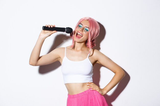 Portrait de joyeuse belle femme en perruque rose, avec un maquillage lumineux, chant karaoké dans le microphone, debout.