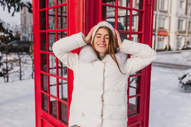 Portrait joyeuse étonnante jeune femme dans des vêtements blancs chauds se détendre sur le soleil en matin gelé en hiver sur la cabine téléphonique rouge