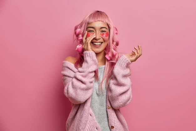 Portrait de joyeuse fille asiatique utilise des patchs d'hydrogel avec effet anti-rides, porte des bigoudis sur les cheveux roses, sourit sincèrement, porte un pull décontracté