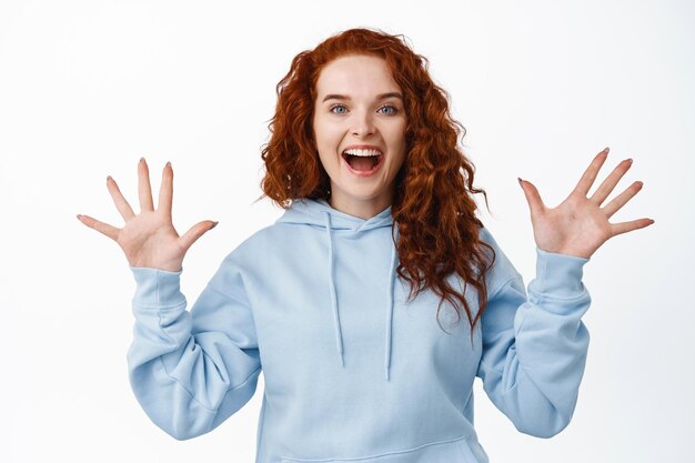 Portrait d'une joyeuse fille au gingembre avec une coiffure frisée, levant les mains et criant de joie, criant de joie et d'émotions positives, debout sur blanc