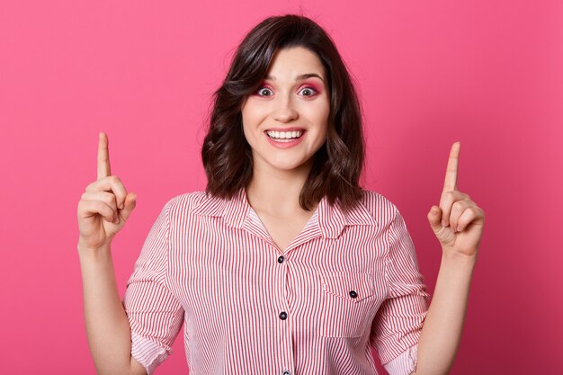 Portrait de joyeuse fille portant une chemise rayée, regardant directement la caméra, pointant les deux index vers le haut, jeune femme avec une excellente idée, debout contre le mur de roses.