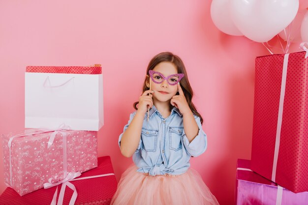 Portrait joyeuse jeune fille en chemise bleue tenant un masque sur le visage autour de coffrets colorés sur fond rose. Beaux moments doux de petite princesse, enfant assez sympathique