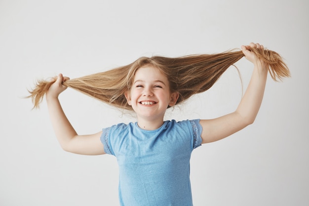 Portrait de joyeuse petite fille blonde mignonne en t-shirt bleu, rire avec les yeux fermés, tenant les cheveux avec les mains, s'amuser.