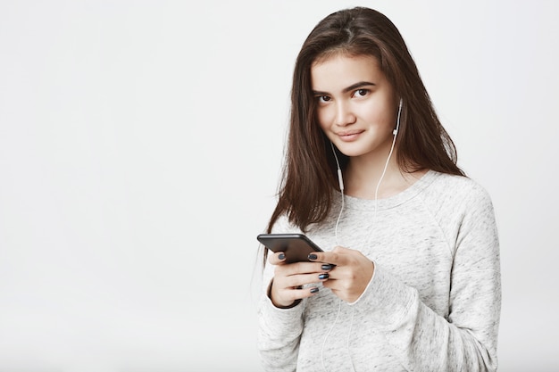 Portrait de joyeux beau modèle européen aux longs cheveux bruns, tenant le smartphone tout en souriant largement et en écoutant de la musique