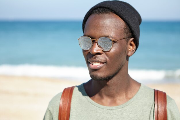 Portrait de joyeux homme noir voyageur profitant des vacances d'été au bord de la mer, à la recherche insouciante et détendue, portant un chapeau à la mode et des lunettes de soleil à lentille miroir. Tourisme, voyages, personnes et style de vie