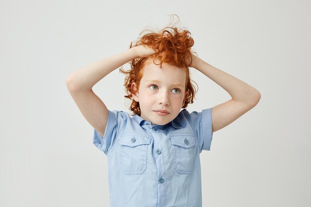 Portrait de joyeux petit garçon au gingembre avec des cheveux ondulés et des taches de rousseur tenant les cheveux dans les mains, regardant de côté avec une expression fatiguée et ennuyée.