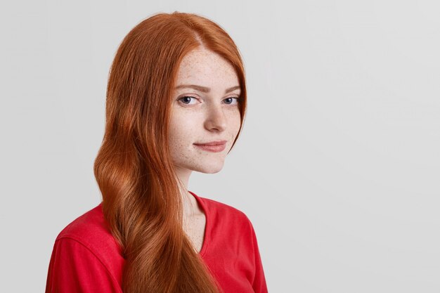 Portrait latéral du modèle féminin aux cheveux roux grave avec taches de rousseur regarde en toute confiance dans l'appareil photo, pose sur blanc avec copie espace pour votre texte promotionnel. Femme gingembre confiante.