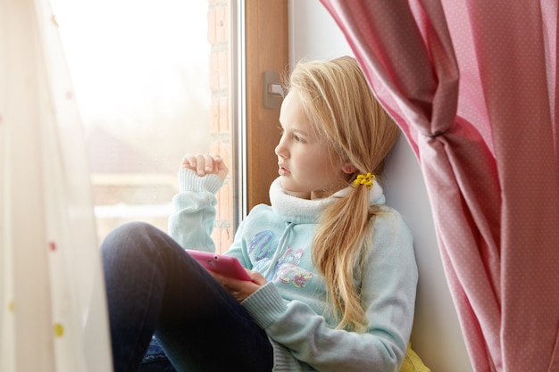 Portrait latéral intérieur de belle enfant de sexe féminin aux cheveux blonds se détendre à la maison sur le rebord de la fenêtre