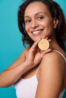 Portrait latéral d'une jolie femme ravie tenant une moitié de citron jaune frais juteux près de son visage et souriant avec un beau sourire à pleines dents en bonne santé posant sur fond bleu clair copier l'espace publicitaire