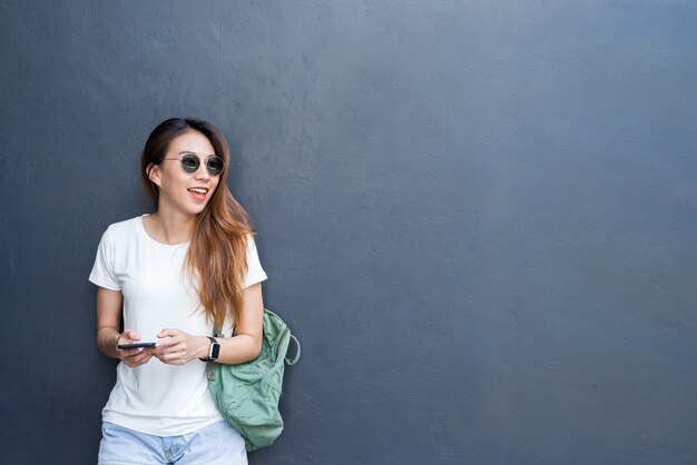 Portrait lifestyle en plein air de jolie jeune fille asiatique sexy dans le style de voyage et de lunettes sur mur gris