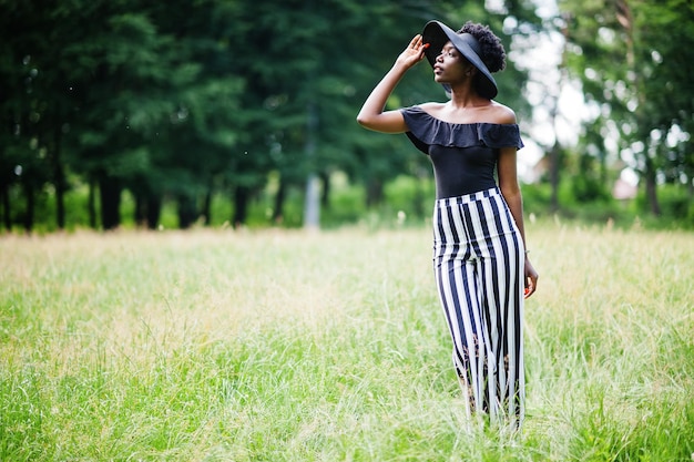 Portrait d'une magnifique femme afro-américaine de 20 ans portant un pantalon rayé noir et blanc et un chapeau d'été posant sur l'herbe verte dans le parc