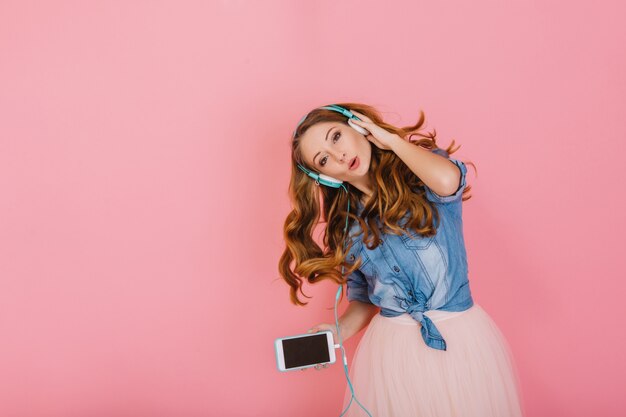 Portrait de magnifique fille aux cheveux longs heureuse dans les écouteurs chantant la chanson préférée isolée sur fond rose. Jolie jeune femme bouclée en chemise en jean avec smartphone dansant et appréciant la musique