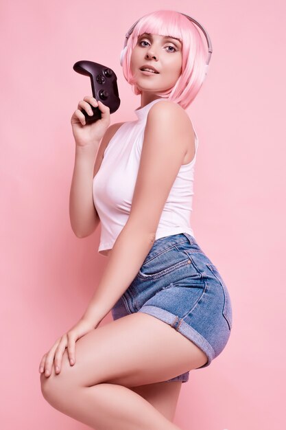 Portrait de magnifique fille heureuse gamer aux cheveux roses, jouer à des jeux vidéo à l'aide de joystick sur coloré en studio