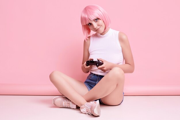 Portrait de magnifique fille heureuse gamer aux cheveux roses, jouer à des jeux vidéo à l'aide de joystick sur coloré en studio