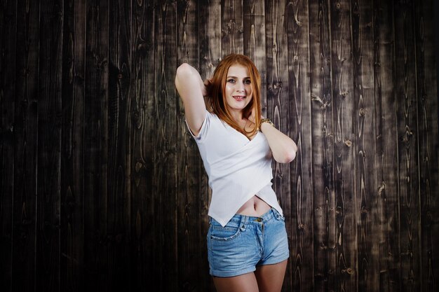 Portrait d'une magnifique fille rousse en t-shirt blanc et short en jean posant dans le studio à côté du mur en bois