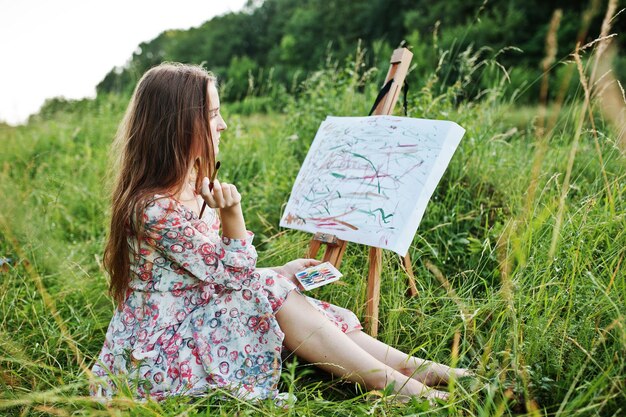 Portrait d'une magnifique jeune femme heureuse en belle robe assise sur l'herbe et peinture sur papier à l'aquarelle