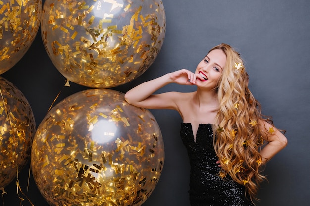 Portrait magnifique jeune femme ludique avec de longs cheveux blonds bouclés s'amusant avec de gros ballons pleins de guirlandes d'or sur l'espace noir