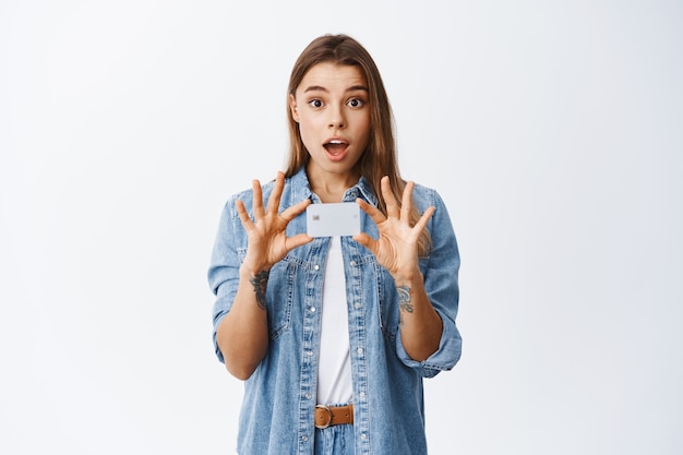 Le portrait d'une magnifique jeune femme en vêtements décontractés présente une publicité bancaire, montrant une carte de crédit en plastique et regardant émerveillé devant le mur blanc